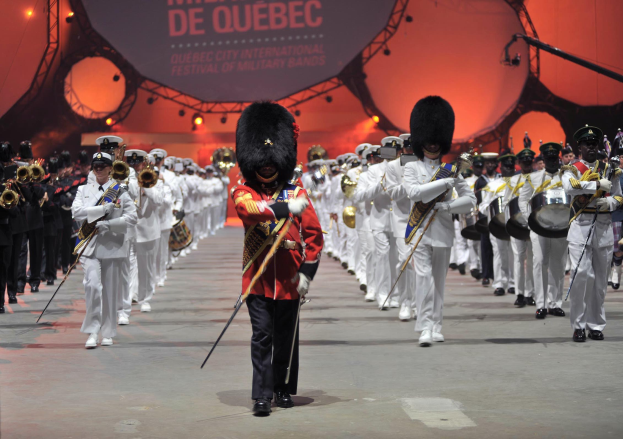Eine Gruppe uniformierter Menschen marschiert auf einer Straße, einige halten Musikinstrumente, mit einer beleuchteten Tafel im Hintergrund, auf der 'Montreal International Festival of Military Bands' steht.