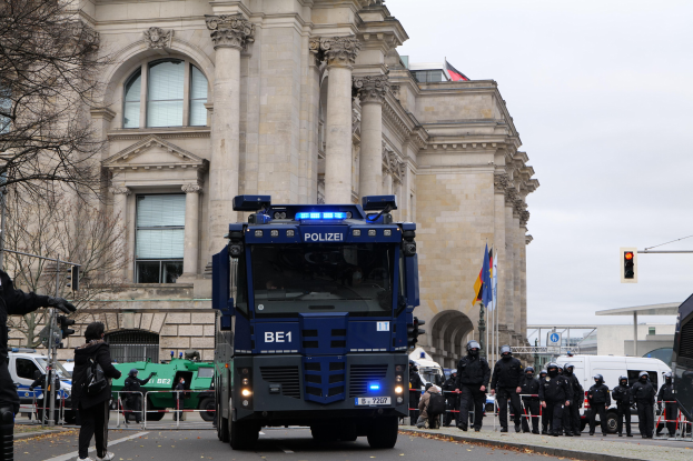 Polizeibeamte stehen vor einem großen Gebäude mit Fenstern, Säulen und Bögen, mit Fahrzeugen auf der Straße und einer Person mit einer Kamera links; Bäume, Verkehrsampeln, Fahnenmasten und ein klarer blauer Himmel sind im Hintergrund zu sehen.