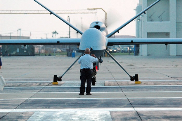 RQ-1 Predator Unmanned Aerial Vehicle auf dem Flughafenvorfeld mit zwei Personen in der Nähe, vor Buildings, Bäumen, Masten und einem klaren blauen Himmel.