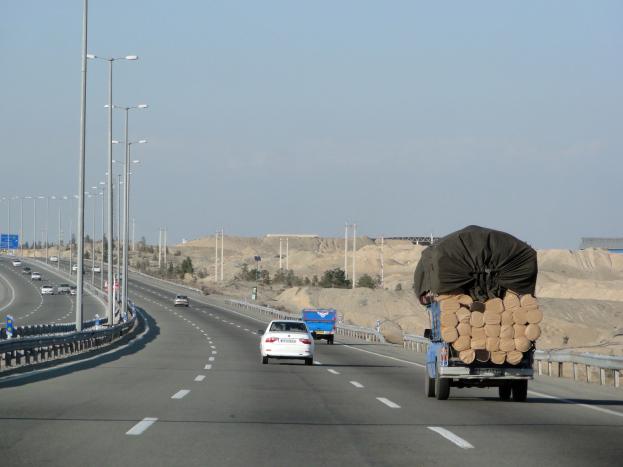 Ein Holzlaster mit einer großen Ladung Holz fährt auf einer Autobahn mit Schutzplanken, Laternenmasten, Schildern, Bäumen und Sand, mit Hügeln und einem klaren blauen Himmel im Hintergrund.