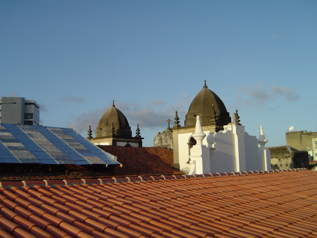Stadtpanorama mit Gebäuden im Vordergrund und einem blauen Himmel im Hintergrund, das Solarpanels auf einem Dach zeigt und damit den Einsatz erneuerbarer Energien anzeigt.
