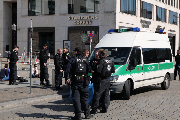 Eine Gruppe von Polizeibeamten steht vor einem Starbucks-Café, rechts ein Van und links einige Menschen, im Hintergrund ein Gebäude mit Fenstern, einem Schild, einem Laternenpfahl und einem Zaun.