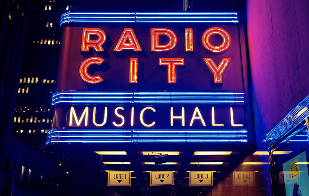 Hell erleuchtetes Radio City Music Hall in New York City mit hohen umliegenden Gebäuden, das eine "Radio City"-Leuchtschrift und zusätzliche Schilder auf der rechten Seite zeigt.