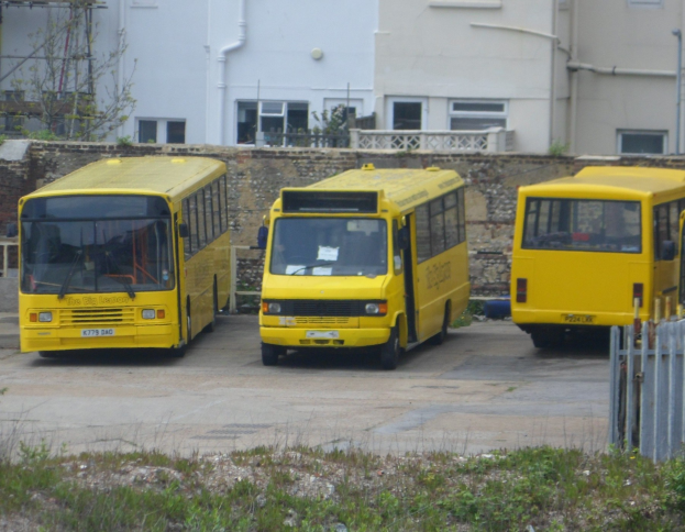 Drei gelbe Busse nebeneinander auf einem eingezäunten Parkplatz mit Grünfläche, einem Gebäude und anderen Strukturen geparkt