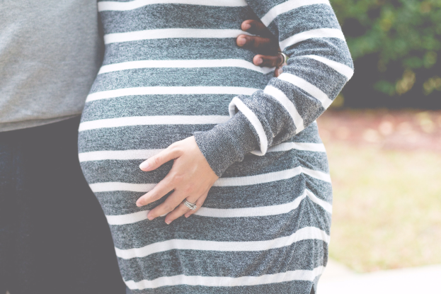 Schwangeres Paar, das Hand in Hand vor einem Wald steht und seinen Babybauch streichelt.