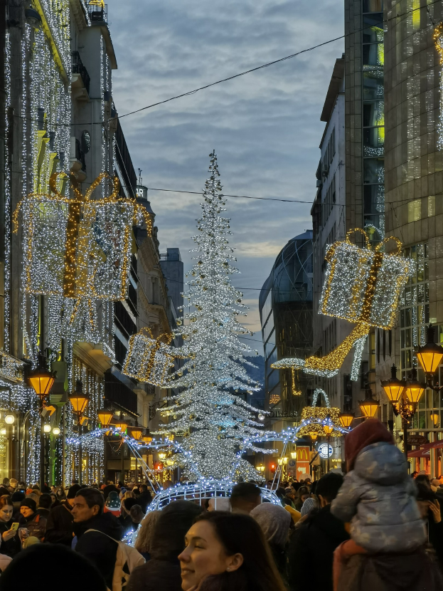 Eine belebte Stadtstraße bei Nacht, geschmückt mit festlichen Weihnachtslichtern, voller Fußgänger und mit einem großen Weihnachtsbaum in der Mitte der Straße.