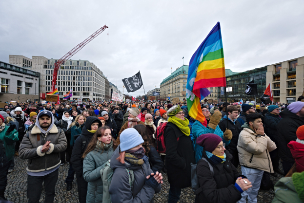 Große Gruppe von Menschen bei einer LGBTQ+-Rechtsdemo in Berlin, die Fahnen und Plakate schwenken, mit Gebäuden, einem Kran und Wolken im Hintergrund.