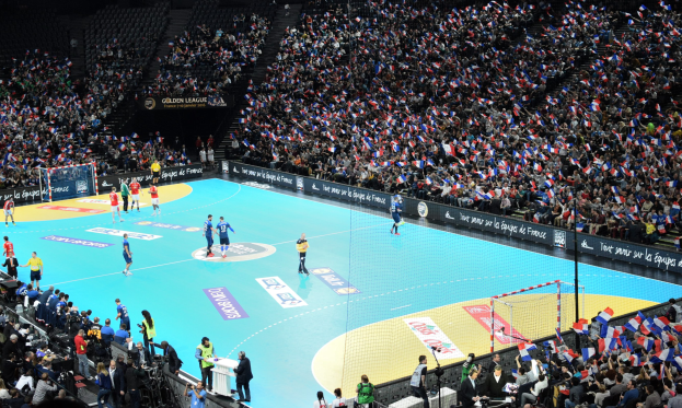 Eine Gruppe von Menschen, die Handball in einem Stadion mit einem Netz auf dem Platz und Zuschauern in den Rängen spielen, während Fotografen das Spiel festhalten.