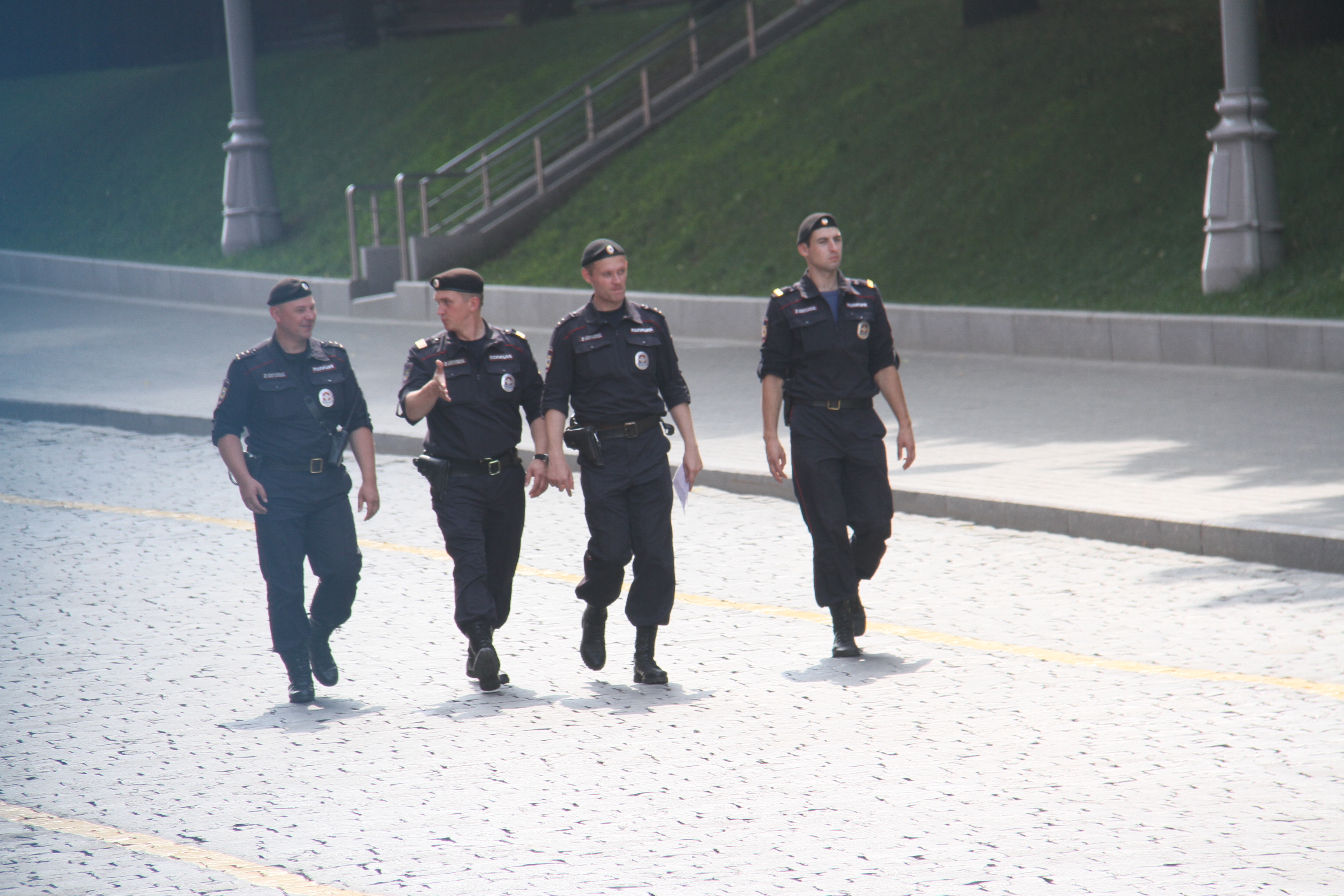 Drei Polizisten in Uniform, die eine Straße entlanggehen, mit Pfählen, Geländern, Treppen und Gras im Hintergrund.