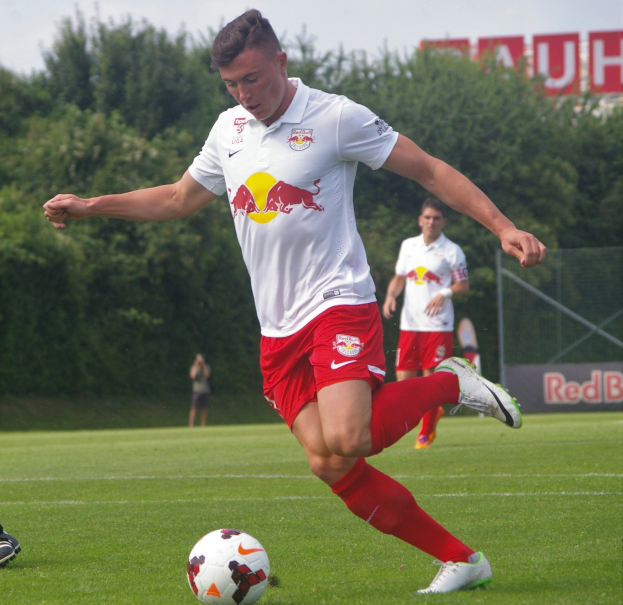 Ein professioneller Fußballspieler in rot-weißer Uniform kickt einen Fußball auf einem grünen Feld mit Bäumen und einem klaren blauen Himmel im Hintergrund, mit "RB Leipzig" auf einem Schild.