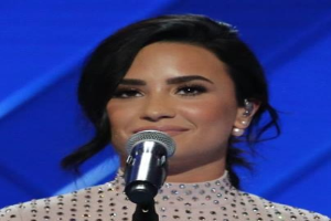 Demi Lovato steht mit einem Mikrofon auf der Bühne der Grammys, tr├Ągt ein schwarzes Kleid mit einem glatten Bob gegen einen blauen Hintergrund, mit einem selbstbewussten Ausdruck.