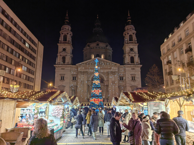 Ein geschäftiger Weihnachtsmarkt vor einer Kirche bei Nacht, mit Menschen um dekorierte Stände versammelt, Gebäuden im Hintergrund und einem sternenklaren Himmel.