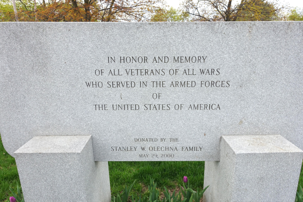 Ein Gedenkstein mit einer Inschrift für US-Streitkräfte-Veteranen, umgeben von Pflanzen, Blumen, Gras, Bäumen und einem bewölkten Himmel.