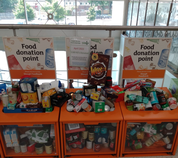 Orange Schübe in einem Gebäude voller gespendeter Lebensmittel wie Flaschen, Dosen und Kisten, mit Informationsschildern und einem Blick auf Fahrzeuge, Bäume, Gebäude und Himmel durch Glasfenster.