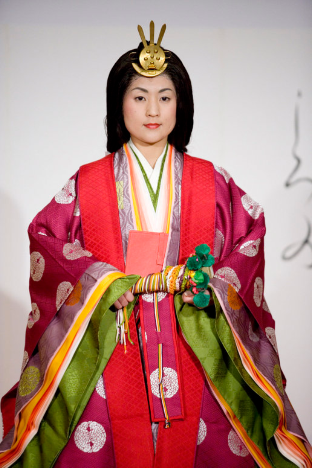 Eine Frau in einem traditionellen japanischen Kimono, das ein leuchtend rotes und grünes Kleid mit aufwändigen Mustern ziert, steht selbstbewusst auf einem Laufsteg mit einem ordentlichen Dutt.