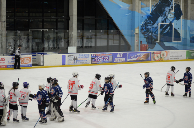 Gruppe junger Menschen, die Eis Hockey auf einer Indoor-Eisbahn spielen, mit Helmen und Stöcken, und einer Person, die links steht und Bannern, Säulen und Glaswänden im Hintergrund.