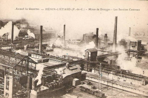 Ein altes Schwarz-Weiß-Foto einer Fabrik mit Rauch, der aus ihren Schornsteinen aufsteigt, umgeben von Gebäuden, Pfählen und Bahnschienen, mit dem Text "Avant la Guerre - Hénin-Lietard - Mines de Douges - Les Services Centraux" oben und unten.