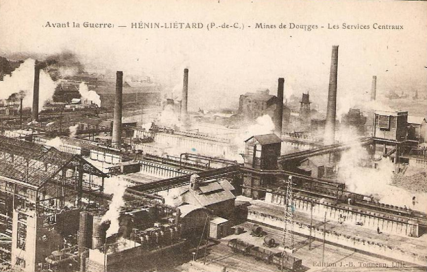 Ein altes Schwarz-Weiß-Foto einer Fabrik mit Rauch, der aus ihren Schornsteinen aufsteigt, umgeben von Gebäuden, Pfählen und Bahnschienen, mit dem Text "Avant la Guerre - Hénin-Lietard - Mines de Douges - Les Services Centraux" oben und unten.