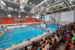 Großer Hallenbad mit vielen Menschen auf Stühlen um das Becken herum, einige im Wasser stehend und Hintergrundtafeln mit Text unter einer Decke mit Lampen und Eisenstangen.