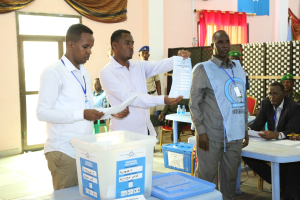 Eine Gruppe von Männern um einen Tisch mit verschiedenen Gegenständen, die bei einer Wahlurne abstimmen, mit Menschen und Möbeln im Hintergrund.