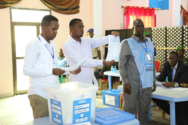 Eine Gruppe von Männern um einen Tisch mit verschiedenen Gegenständen, die bei einer Wahlurne abstimmen, mit Menschen und Möbeln im Hintergrund.
