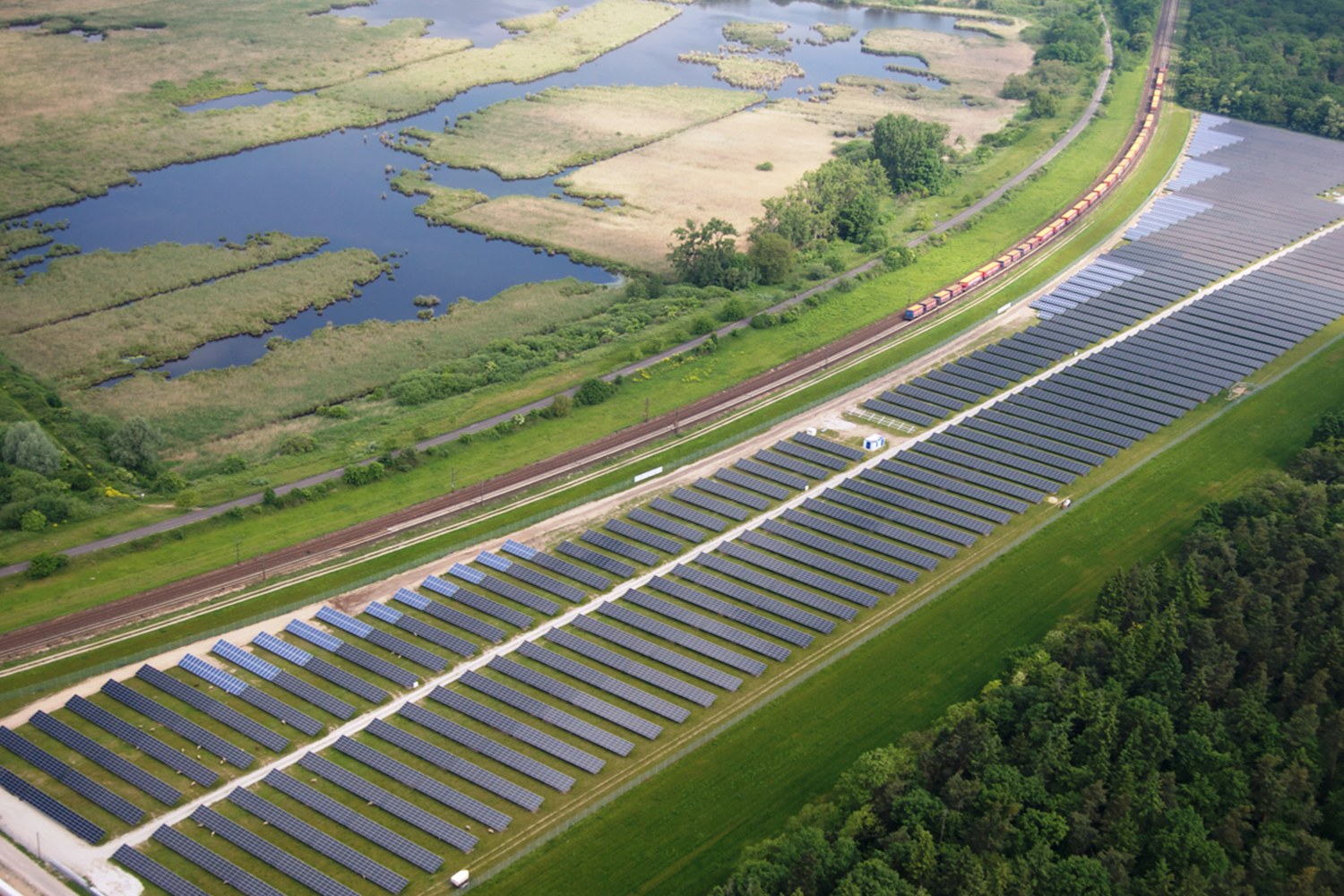 Luftaufnahme eines großen Solarparks in einem Feld mit Bäumen, Gras, Pflanzen und Wasser, mit Solarpanelen und einem Zug auf einer Eisenbahnschiene.