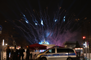 Ein Polizeiwagen steht vor einer Menschenmenge, umgeben von Fahrrädern, Zelten, Laternenmasten, Schildern, Verkehrszeichen, Bäumen, Gebäuden und einer Statue, unter einem von Feuerwerk erhellten Himmel.