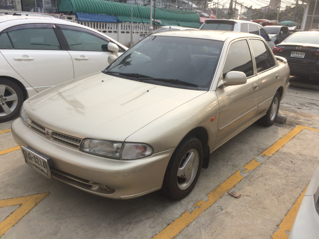 Ein goldfarbener Wagen auf einem Parkplatz neben anderen Fahrzeugen geparkt, mit Gebäuden, Strommästen und Bäumen unter einem klaren blauen Himmel.