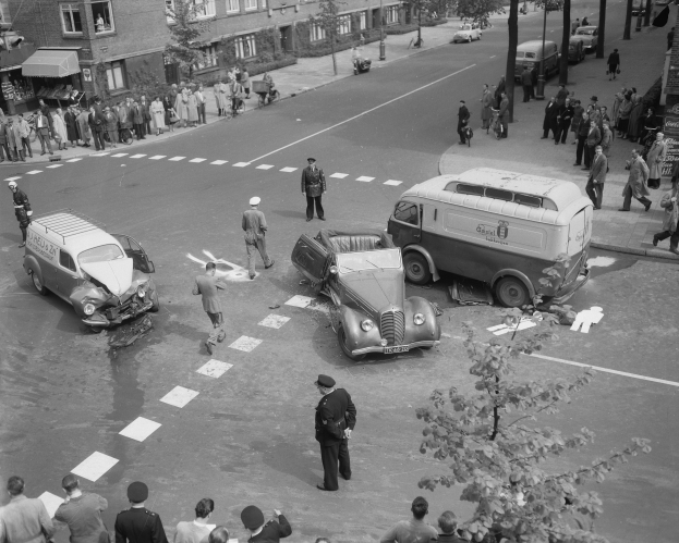 Eine Schwarz-Weiß-Szene eines Autounfalls an einer Straßenecke mit mehreren Fahrzeugen, darunter ein Van, und einer Gruppe von Menschen, die darum herumstehen, mit Gebäuden, Bäumen, Masten und Texttafeln im Hintergrund.