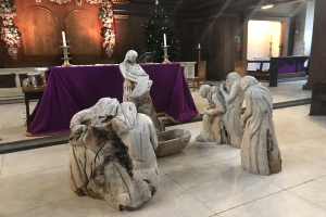 Krippe in einer Kirche mit Statuen auf dem Boden, einem Tisch mit einem Tuch und Kerzenständern, Stühlen, einem geschmückten Weihnachtsbaum und einer Wand im Hintergrund.