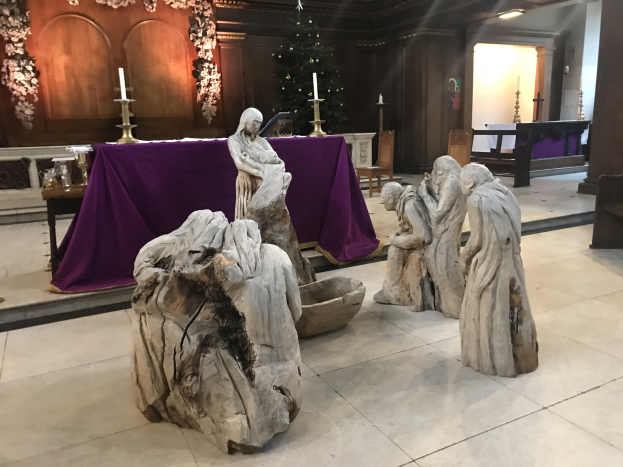 Krippe in einer Kirche mit Statuen auf dem Boden, einem Tisch mit einem Tuch und Kerzenständern, Stühlen, einem geschmückten Weihnachtsbaum und einer Wand im Hintergrund.