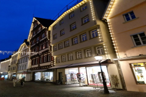 Eine Kopfsteinpflasterstraße in Rothenburg ob der Tauber, Deutschland, geschmückt mit festlichen Weihnachtslichtern, mit Menschen, die spazieren gehen, und einem geparkten Fahrrad, flankiert von Gebäuden mit Fenstern und einem sichtbaren Himmel.