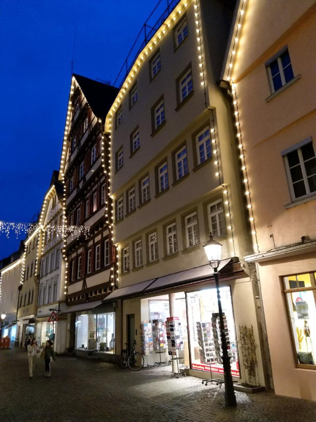 Eine Kopfsteinpflasterstraße in Rothenburg ob der Tauber, Deutschland, geschmückt mit festlichen Weihnachtslichtern, mit Menschen, die spazieren gehen, und einem geparkten Fahrrad, flankiert von Gebäuden mit Fenstern und einem sichtbaren Himmel.