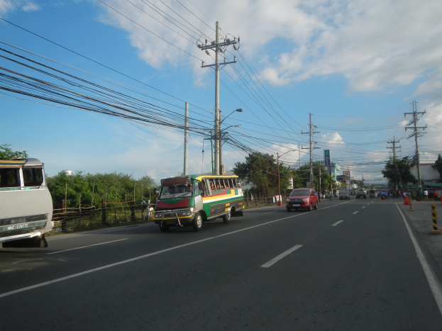 Eine Stadtstraße mit Autos und Bussen, Strommasten mit Drähten, Laternen, Bäumen und einem bewölkten Himmel.