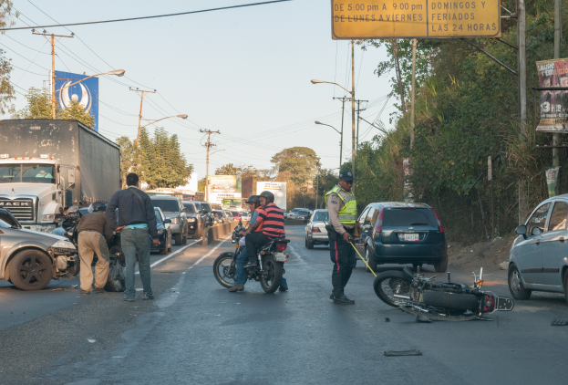 Eine Gruppe von Menschen steht um ein verunglücktes Motorrad auf der Straße mit mehreren Fahrzeugen, darunter ein Lastwagen, und einer Hintergrundlandschaft aus Bäumen, Pfählen, Laternen und Schildern unter dem Himmel.