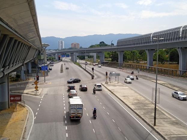 Eine vielbefahrene Autobahn mit mehreren Fahrzeugen in der Nähe einer Brücke, mit Straßenlaternen, Verkehrszeichen, Baumreihen, Gebäuden, Hügeln und einem bewölkten Himmel.