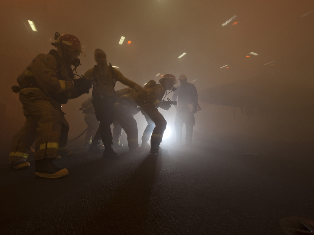 Feuerwehrleute in Helmen gehen öffentlich mit Ausrüstung, mit einem Flugzeug und Lichtern im Hintergrund und einer Straße unten.