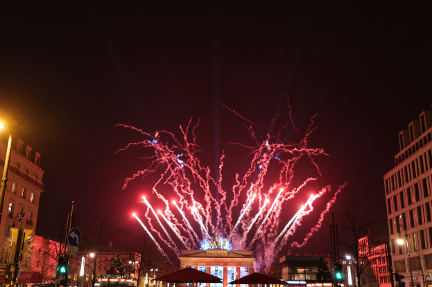 Eine belebte Straßenkreuzung in Berlin an Silvester, voller Menschen, Fahrzeuge und Gebäude, beleuchtet von Feuerwerk und Gebäudelichtern, die eine festliche Atmosphäre schaffen.