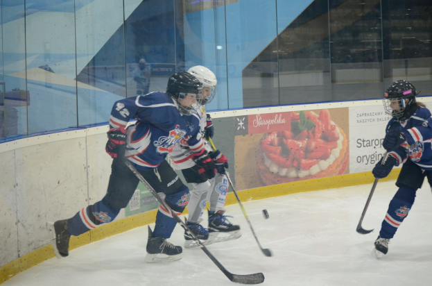 Gruppe junger Menschen, die Eis hockey auf einer Indoor-Eisfläche spielen, mit Helmen, Sportuniformen und Schlittschuhen, während sie Hockey-Schläger halten, mit einer Glaswand und einem Plakat im Hintergrund.