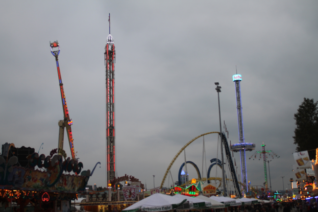 Eine Menschenmenge versammelt sich um einen nächtlichen Fasching, beleuchtet von bunten Lichtern von Fahrgeschäften, Pfählen und Bäumen, unter einem bewölkten Himmel.