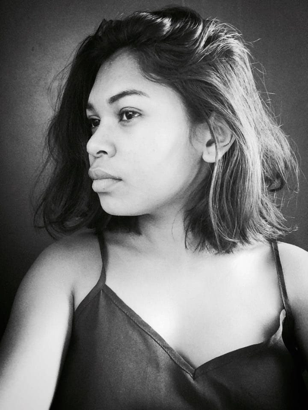 Eine selbstbewusste Frau in einem schwarzen Kleid posiert in einem Schwarz-Weiß-Foto vor einer Wand, ihr glatter Bob und das dezente Make-up unterstreichen ihre selbstbewusste Haltung.