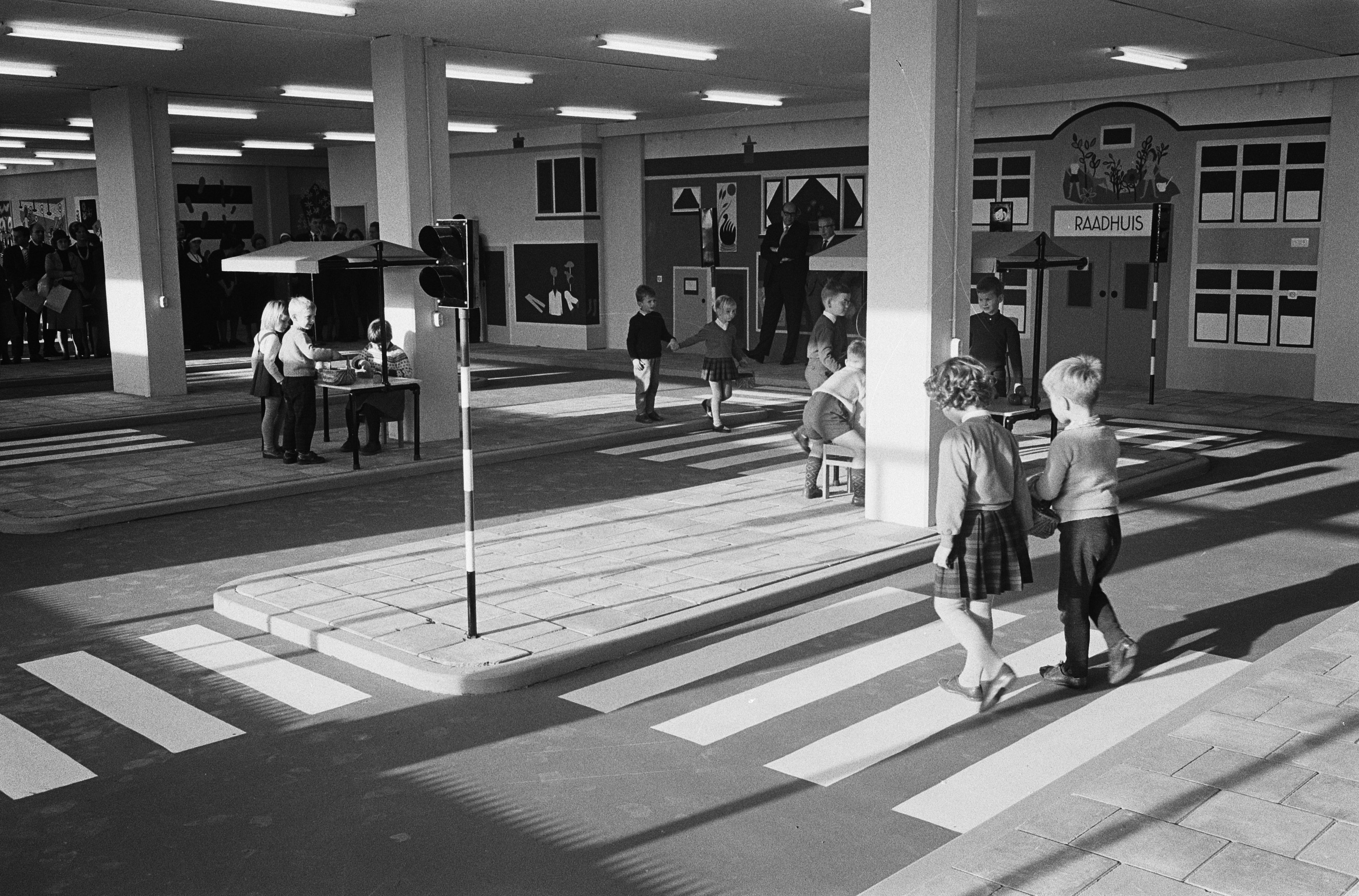 Schwarz-weißes Foto von Kindern, die eine Straße an einer Ampel überqueren, mit Fußgängern in der Nähe, einem Verkehrssignalmast im Vordergrund und strukturellen Säulen und Leuchten im Hintergrund.