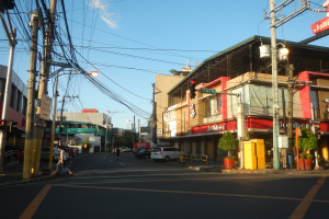 Eine Stadtstraße mit Autos, Fußgängern, Strommasten, Verkehrszeichen, Gebäuden mit Schildern, Topfpflanzen und einer bewölkten Himmel