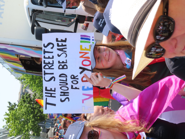 Eine Gruppe von Menschen hält ein Schild mit der Aufschrift "Die Straßen sollten sicher sein" vor einem Lastwagen, einige tragen Mützen und Schutzbrillen und andere halten Schilder, vor einem Hintergrund aus Bäumen, Gebäuden und einem klaren blauen Himmel.