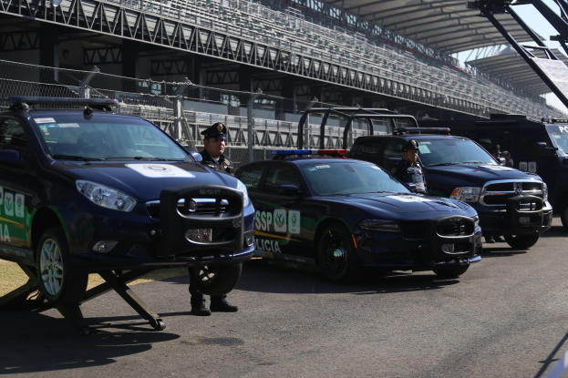 Polizeifahrzeuge vor einer Rennstrecke geparkt, uniformierte Personen auf der Straße, Stadion und Zaun im Hintergrund.