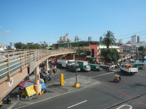 Eine belebte Straße mit starkem Verkehr neben einer Brücke, mit Fahrzeugen, Fußgängern, Zelten, Versorgungsmasten, Verkehrssignalen, Bäumen, Gebäuden und Wasser im Hintergrund unter einem bewölkten Himmel.