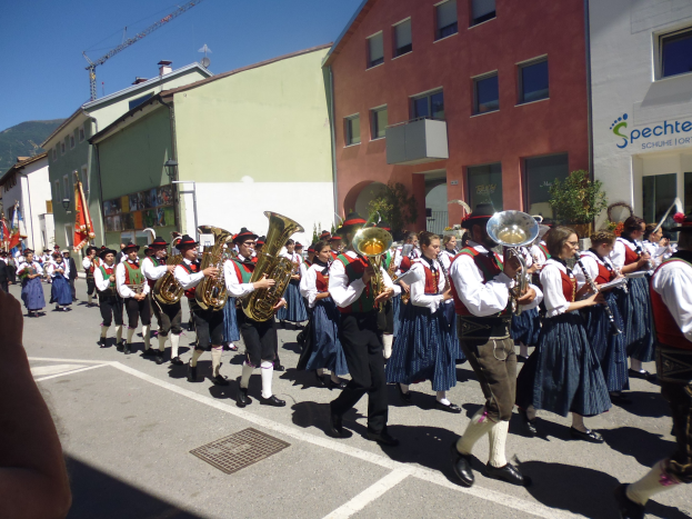 Eine Gruppe von Menschen in traditioneller bayrischer Tracht, die Musikinstrumente spielen, während sie eine Straße mit Gebäuden entlanggehen, einige halten Fahnen, mit einem Hügel und einem klaren blauen Himmel im Hintergrund.