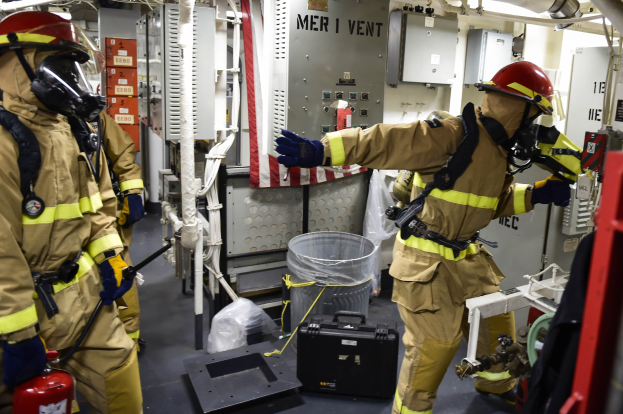 Drei Feuerwehrleute in Schutzausrüstung stehen zusammen in einem unordentlichen Raum mit einem Feuerlöscher links, einer Maschine rechts und Rohren und Lampen oben.