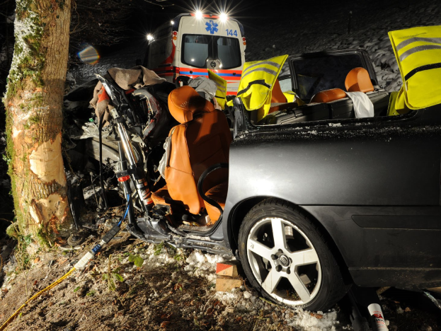 Ein beschädigt auf der Seite einer schneebedeckten Straße stehendes Auto mit einem im Hintergrund sichtbaren Rettungswagen.