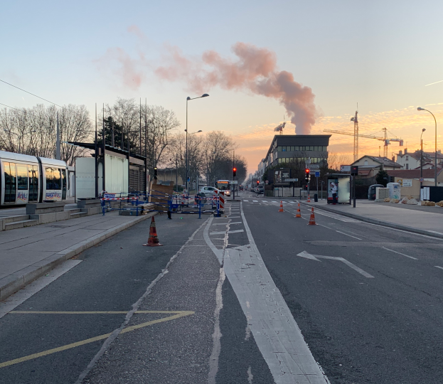 Eine Stadtstraße mit Verkehrskegeln, einem Bus, Laternen, Bäumen, Gebäuden, Kränen und Rauch, der aus einem fernen Gebäude aufklaren Himmel aufsteigt.